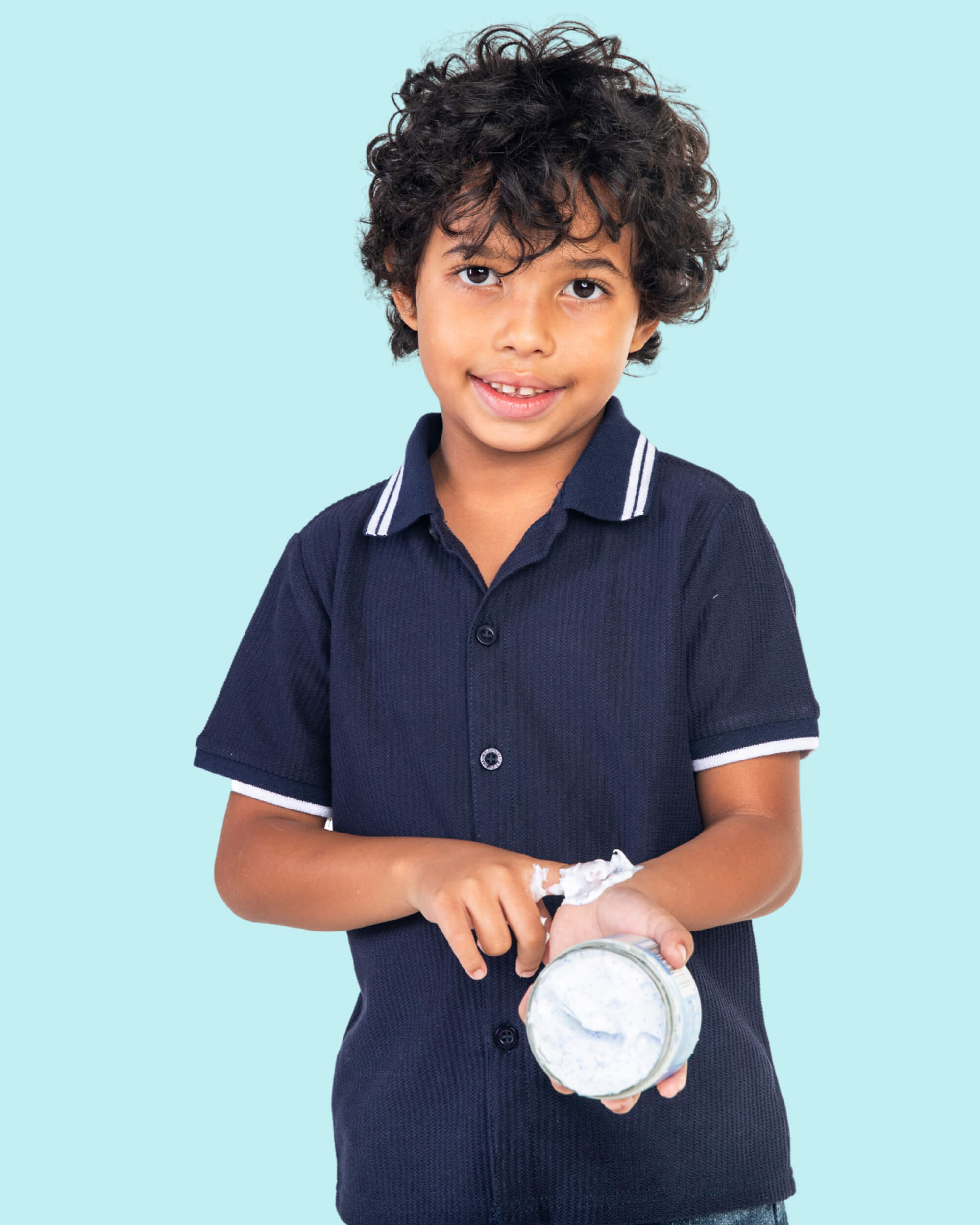 A boy in a navy shirt holding a Tiiglu® Kids Dragon Body Crème jar (150ml) with cream on his wrist, on a light blue background. Fruity bubblegum-scented, &