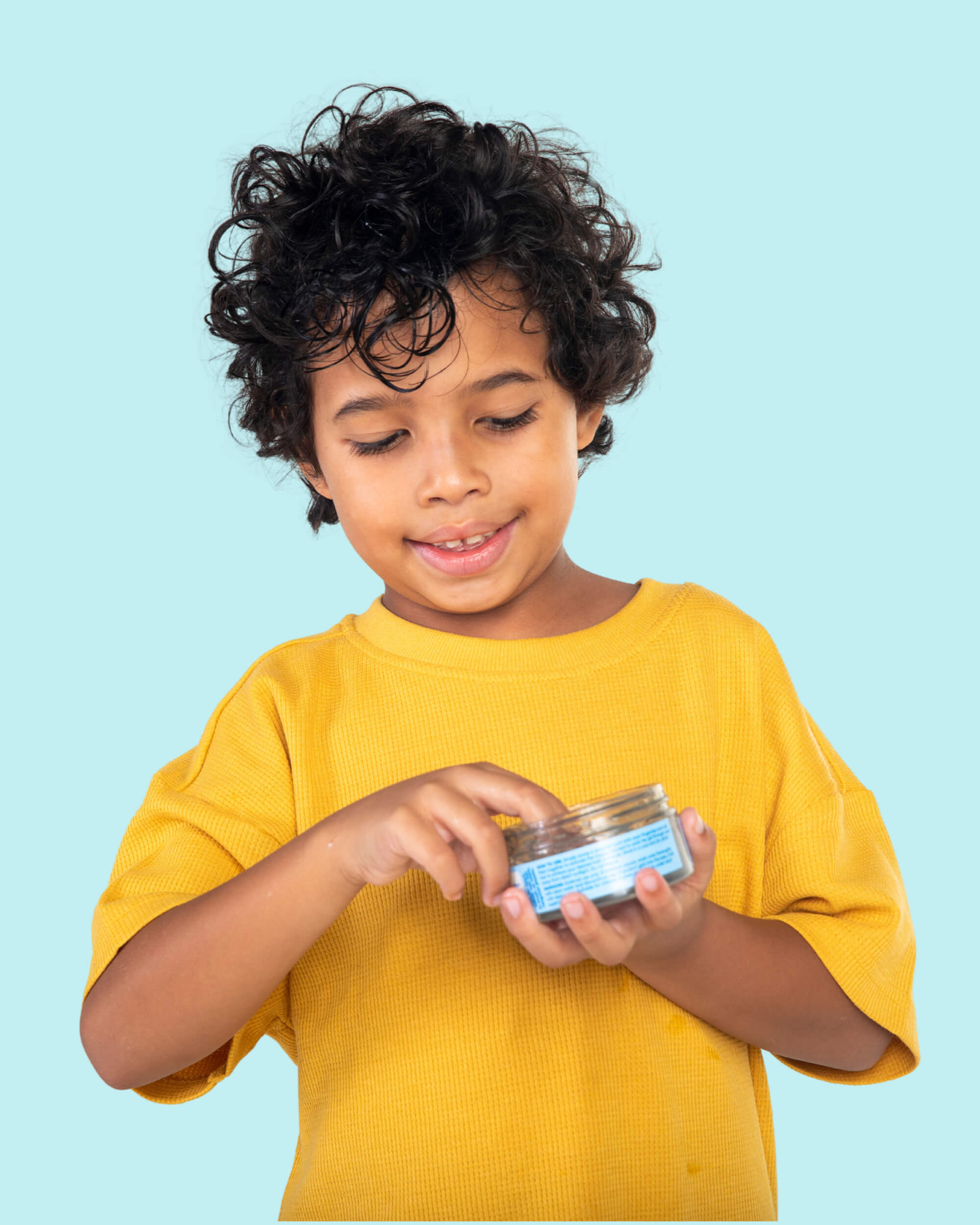 A young boy in a yellow shirt scooping clear, fragrance-free gel from a Tiiglu® Kids Dragon Styling Gel jar (100ml/3.3 fl. oz.) against a light blue background. The jar label features a green dragon and text noting &