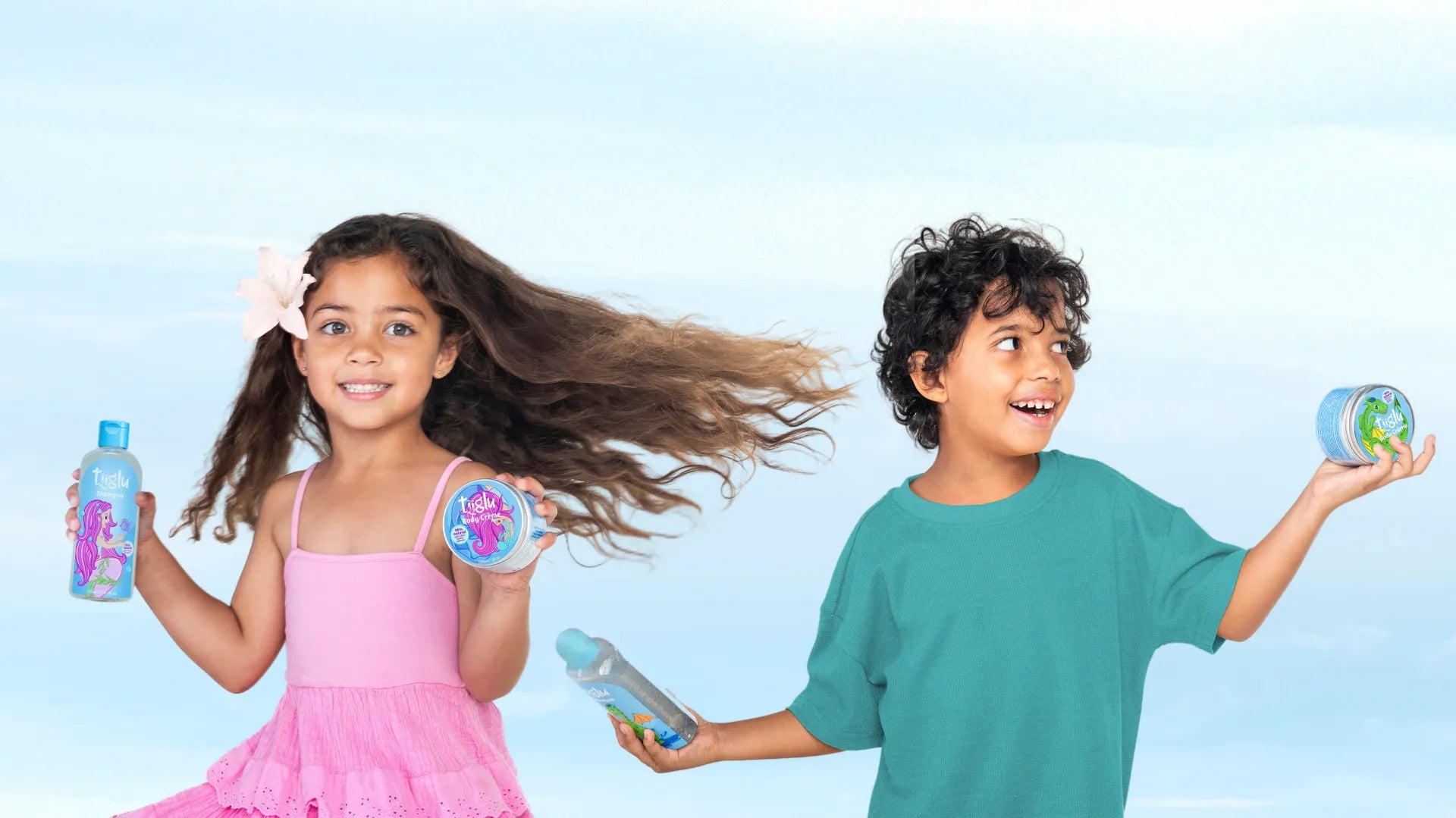 A little girl in a pink dress with a flower in her hair and a boy in a green shirt hold Tiiglu® body wash and crème with mermaid and dragon designs, against a light blue sky. This home banner promotes gentle, nourishing body and hair care for kids aged 3+