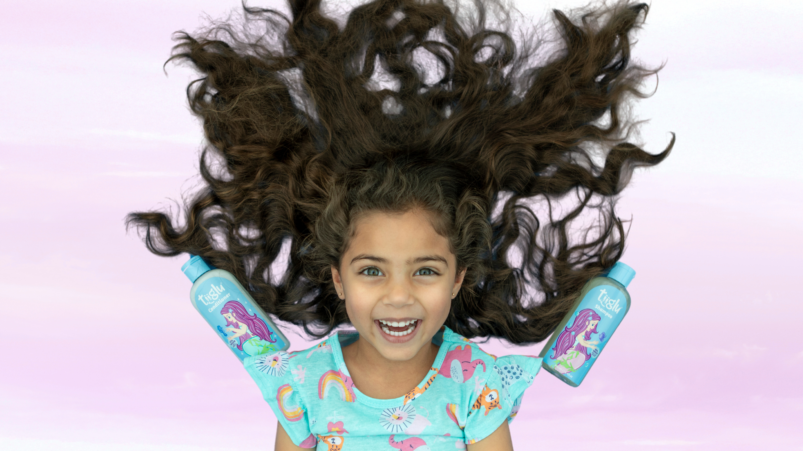 A girl with long, wavy hair lies on her back, smiling, with two Tiiglu® hair care bottles featuring mermaid designs near her shoulders. This Hair section banner promotes gentle shampoos and conditioners with jojoba and coconut oils for kids' hair.