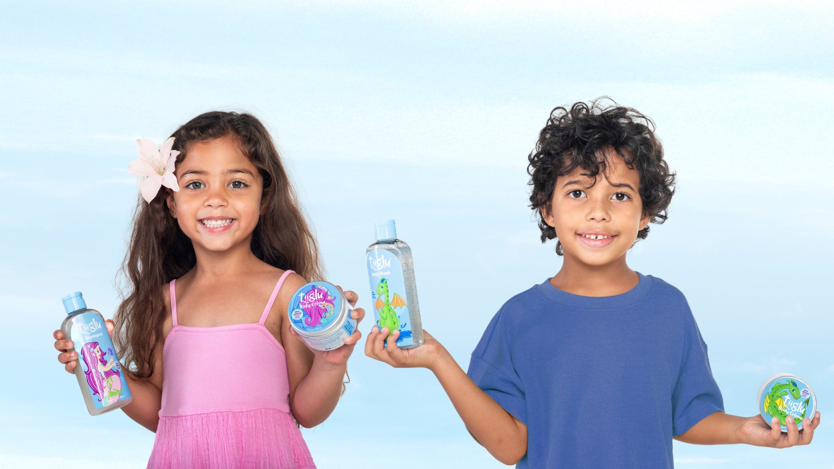  little girl in a pink dress and a boy in a blue shirt hold Tiiglu® body wash and crème with mermaid and dragon designs. This Bath & Body banner promotes playful washes and crèmes with shea butter for kids' skin.