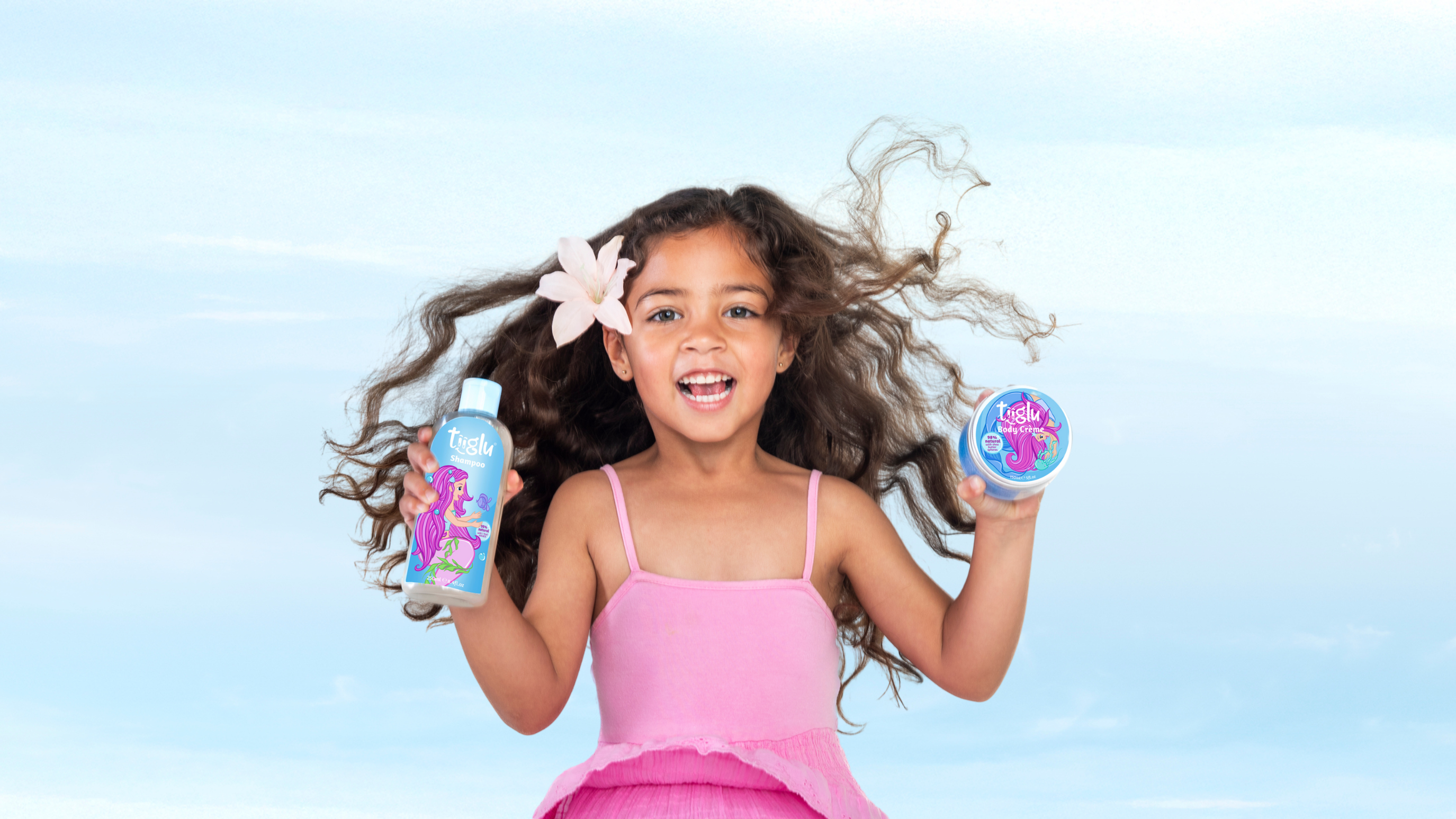 A little girl in a pink dress with a flower in her hair holds a Tiiglu® body wash and crème with mermaid designs. The background is a light blue sky. This Mermaid section banner promotes products to gently cleanse and nourish kids' skin and hair.