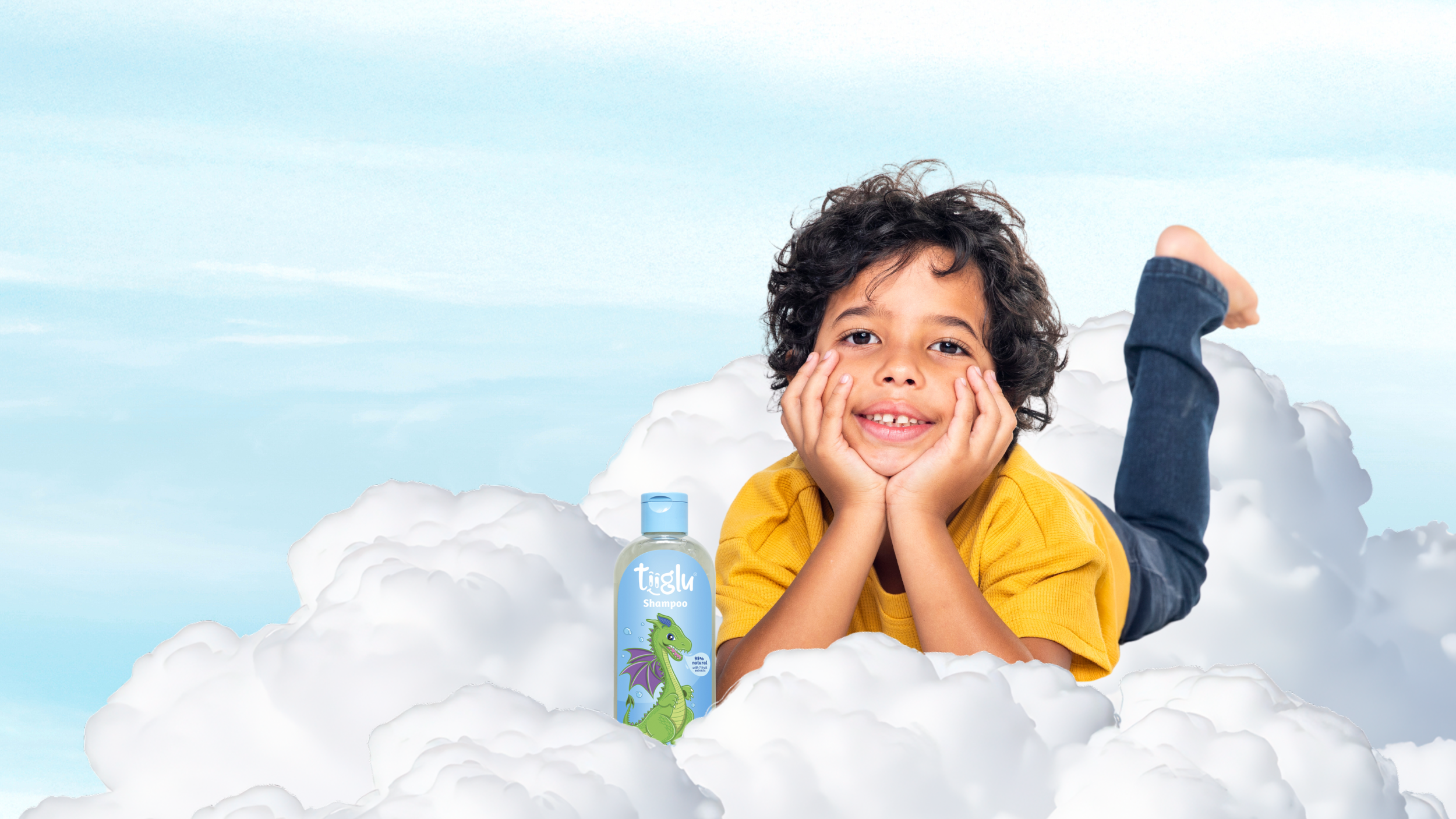 A little boy in a yellow shirt lies on clouds with a Tiiglu® body wash bottle featuring a dragon design. This Dragon range banner promotes fruity fresh body and hair care with shea butter to cleanse kids' skin.