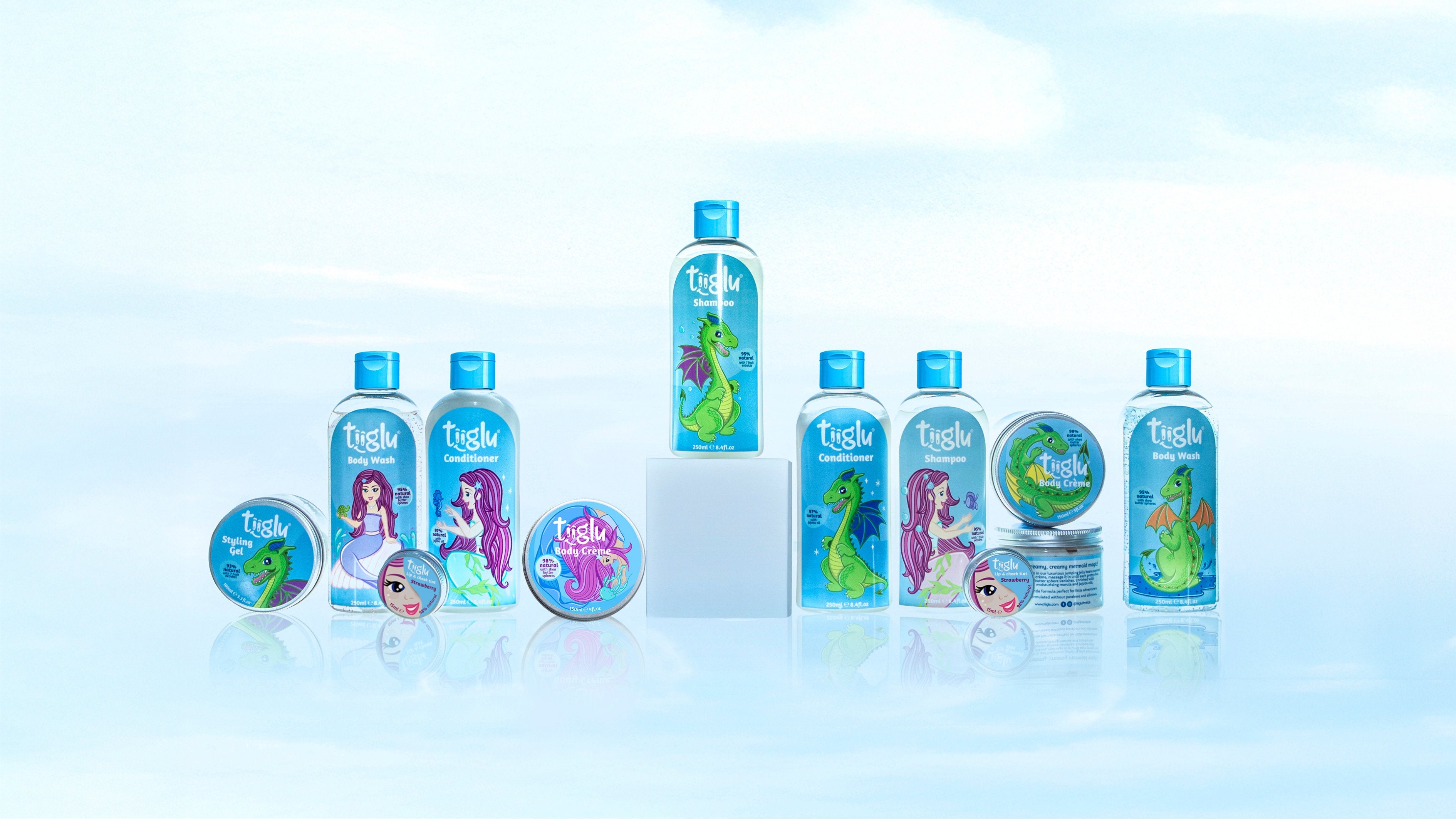 A collection of Tiiglu® body and hair care products with dragon and mermaid designs is displayed on a light blue sky background. This Shop All banner promotes gentle, highly natural products for kids 3+, with shea butter and oils to nourish skin.
