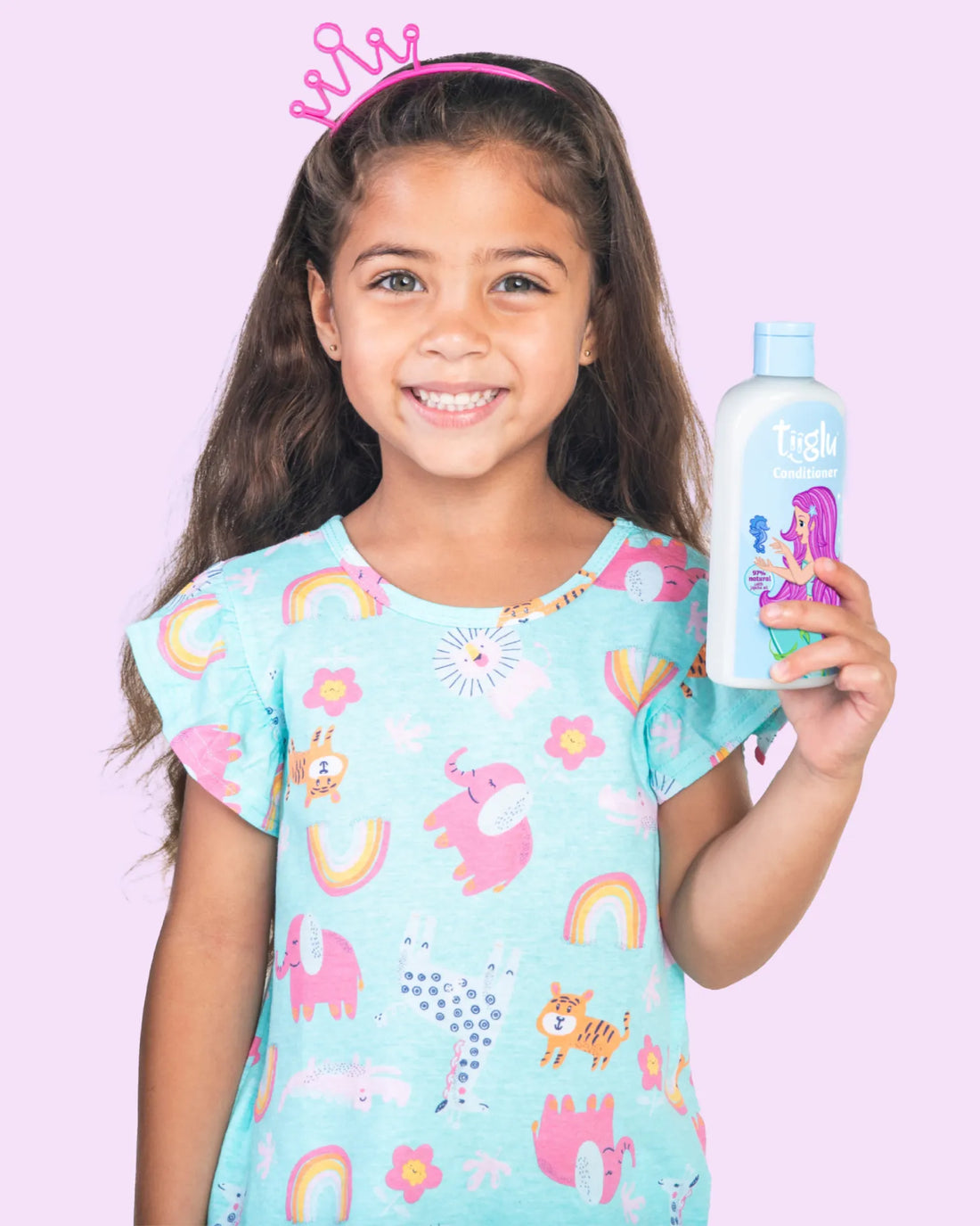 A girl in colourful animal print pyjamas with a pink crown, holding a Tiiglu® Kids Mermaid Conditioner bottle (250ml) with a pink-haired mermaid on a light blue label, &