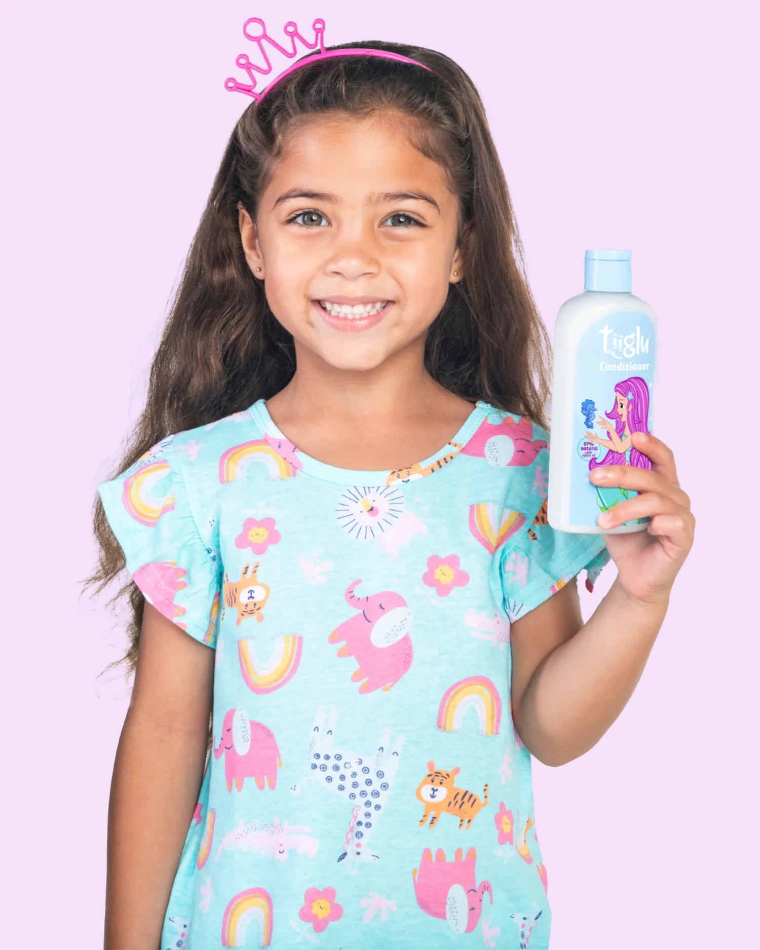 A girl in colourful animal print pyjamas with a pink crown, holding a Tiiglu® Kids Mermaid Conditioner bottle (250ml) with a pink-haired mermaid on a light blue label, &
