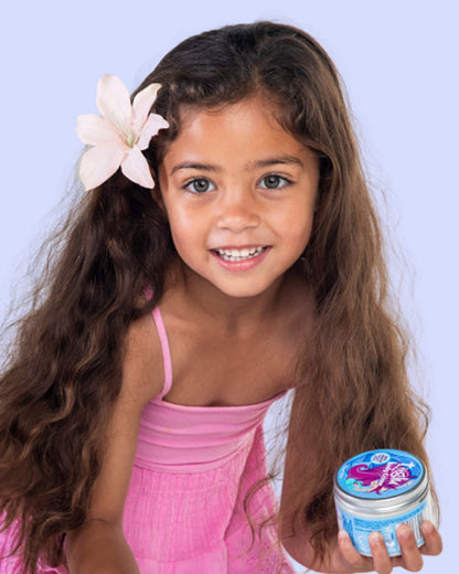 A girl in a pink dress with a pink flower in her hair, holding a Tiiglu® Kids Mermaid Body Crème jar (150ml) with a pink-haired mermaid on a light blue label, &