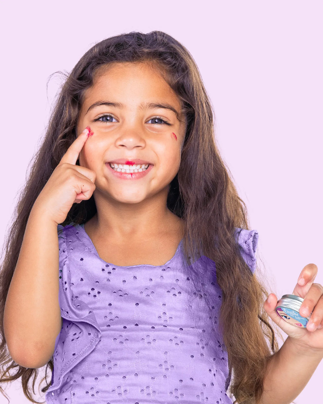 A girl in a purple dress holding a Tiiglu® Kids Mermaid Lip &amp; Cheek Tint jar (15ml) with pink tint on her cheek, on a light pink background. Strawberry-scented, &