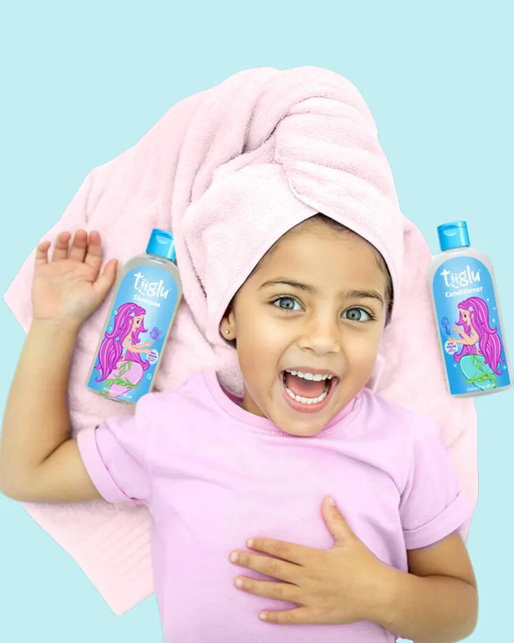 A girl in a pink shirt with a pink towel wrapped around her head lies on her back, holding Tiiglu® shampoo and conditioner bottles with mermaid designs. The Hair section on the home page promotes washing, conditioning, and styling products for kids