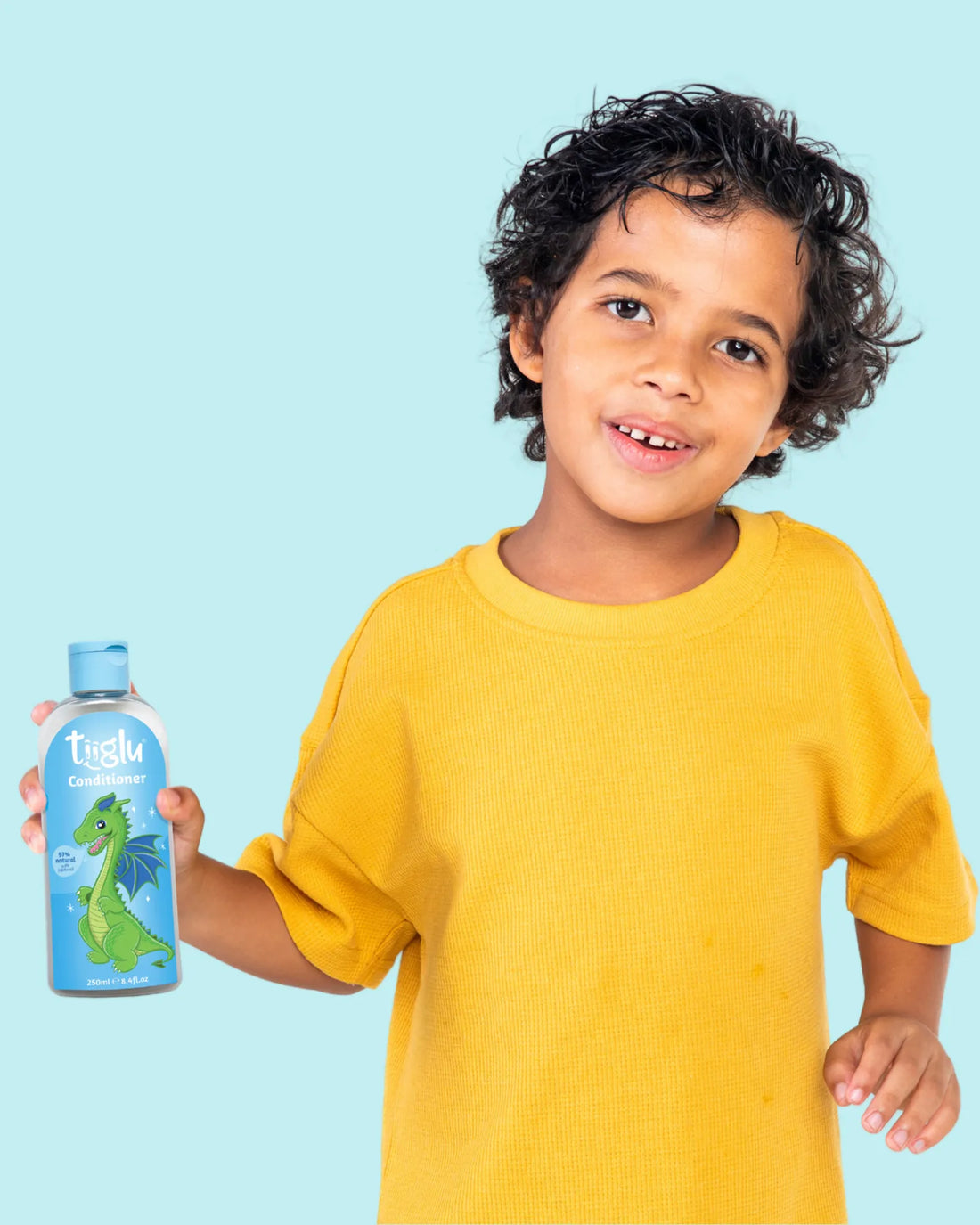 A boy in a yellow shirt holding a Tiiglu® Kids Dragon Conditioner bottle (250ml) with a green dragon on a light blue label, &
